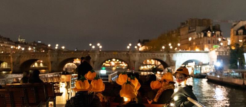 Passagers portant des chapeaux citrouilles sur le bateau, face à un pont illuminé sur la Seine